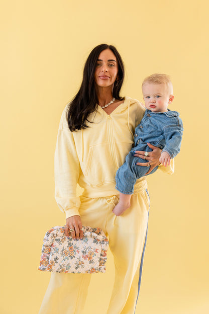 Woman in a yellow set holding a baby and a floral clutch against a yellow background