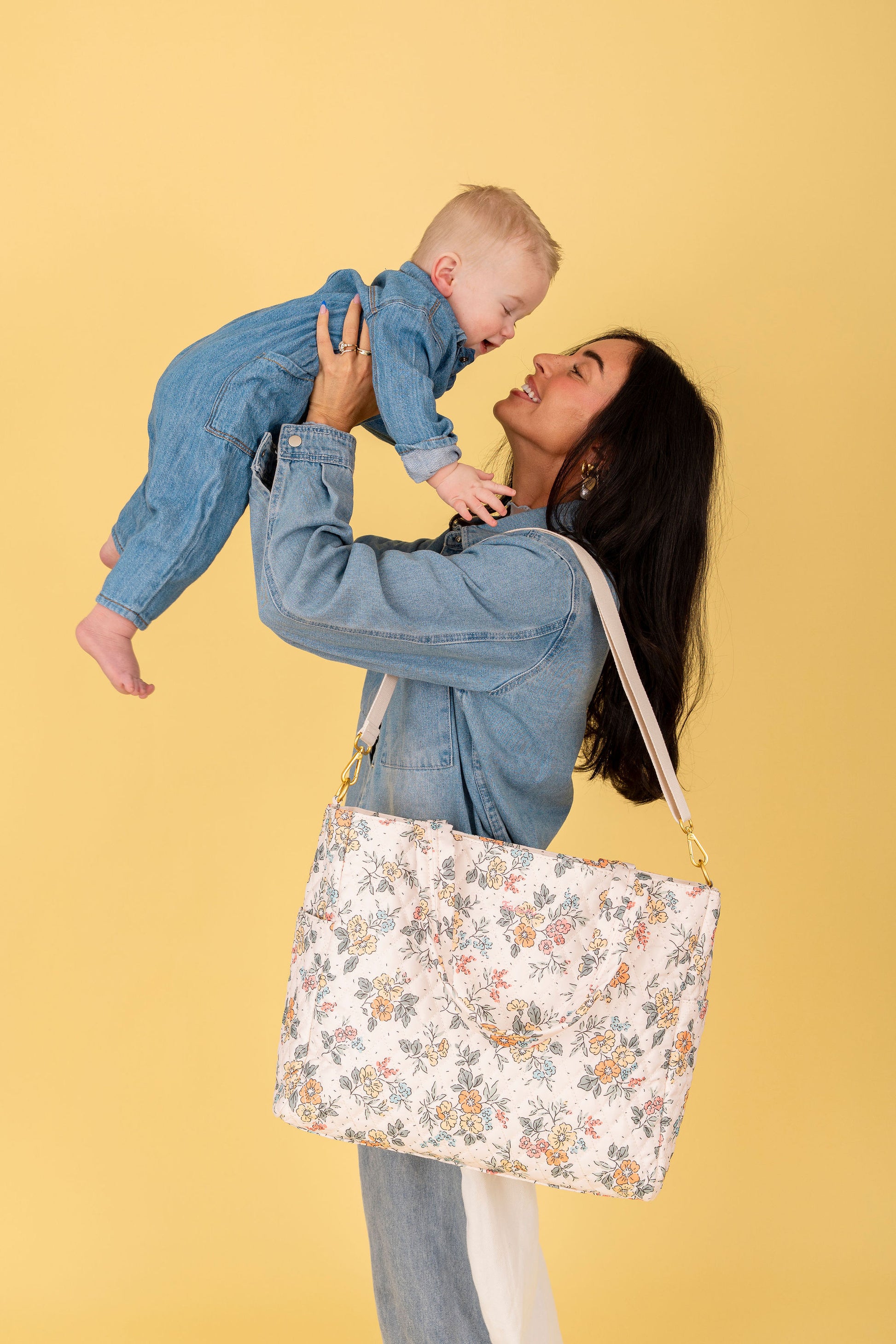Woman holding a baby and a floral tote bag against a yellow background